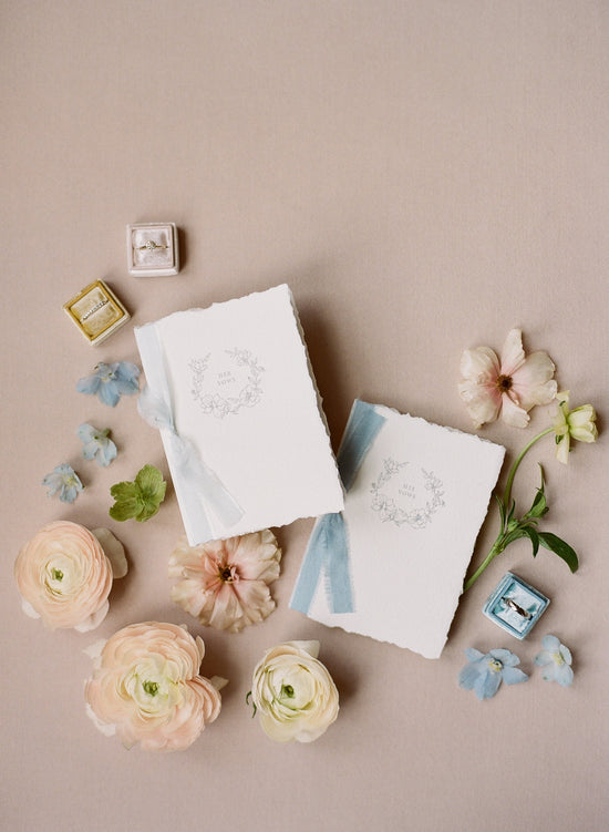 His and her wedding vow books with silk ribbon in flat-lay adorned with flowers and wedding rings