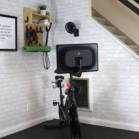 A home gym that has white brick peel and stick wallpaper on the wall behind a peloton bike.