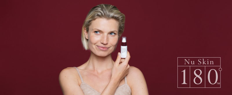 Junge Frau mit blonder Kurzhaarfrisur und straffer Haut hält lächelnd Nu Skin 180º UV Block Hydrator SPF 18 vor einem weinroten Hintergrund neben ihr Gesicht.