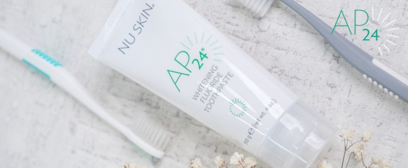 AP 24 Whitening Fluoride Zahnpasta von NuSkin liegt geschlossen auf steinener Oberfläche, daneben eine Zahnbürste.