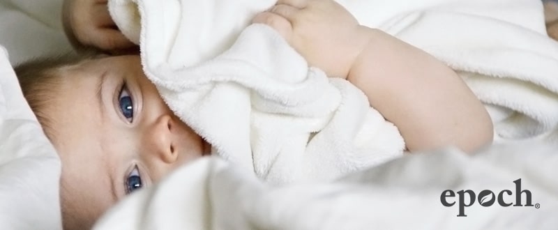 Baby mit blauen Augen eingewickelt in ein weißes flauschiges Handtuch lacht. Das Bild wurde mit dem epoch Logo von NuSkin in der rechten unteren Ecke gebrandet.