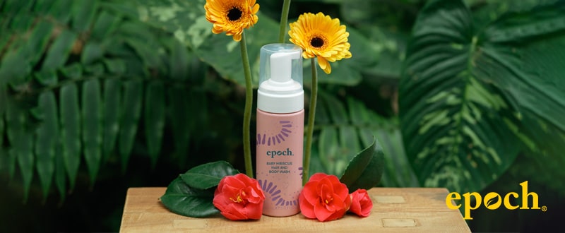 Epoch Baby Hibiscus Hair and Body Wash Flasche steht vor dunkelgrünem Farn auf kleinem Holztisch. Um die Packung wurden Blüten der Hibiscus verteilt. Dahinter stehen Ringelblumen.