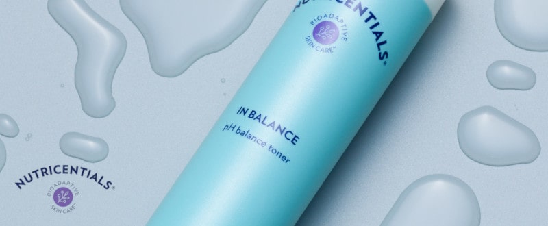 In Balance pH Balance Toner (Nutricentials) Tube (150ml) liegt geschlossen auf glatter hellblauer Oberfläche. Das Produkt ist von großen klaren Wassertropfen umgeben.