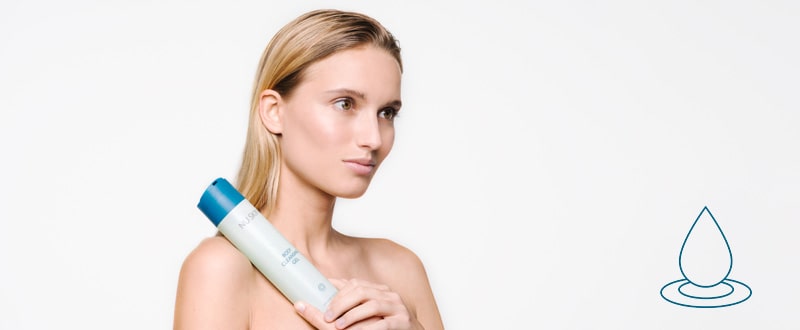 Junge Frau mit blonden frisch gewaschenen Haaren hält die Tube Body Cleansing Gel von Nu Skin in den Händen vor ihren Körper. Sie steht vor einem neutralen weißen Hintergrund.