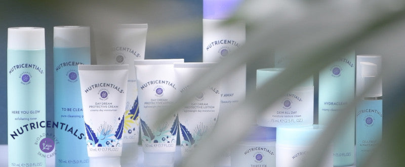 Mehrere Nutricentials Produkte (unter anderem Brighter Day Exfoliant Scrub) stehen aufgereiht in sanitärer Umgebung. Im Vordergrund ragen verschwommen die Blätter einer Pflanze ins Bild.