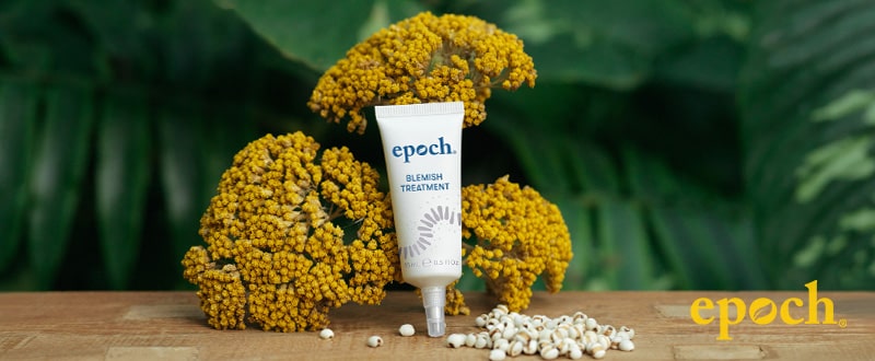 epoch blemish treatment Tube (15ml) steht auf Holztisch umgeben von Scharfgabe-Blüten und dunkelgrünen Blättern.