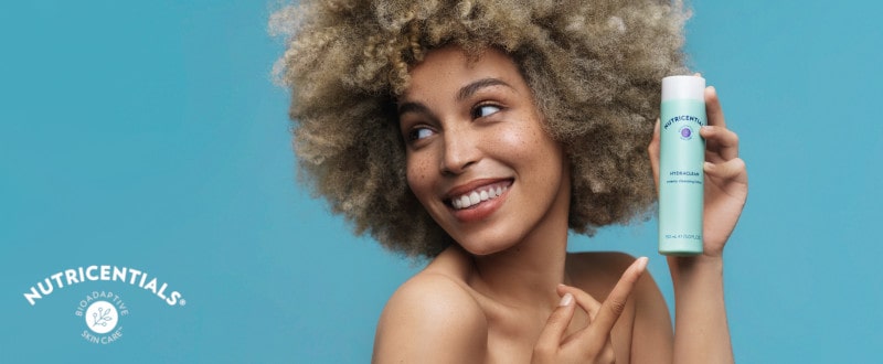 Junge Frau mit Afro und Sommersprossen zeigt breit lächelnd auf die HydraClean Creamy Cleansing Lotion von Nutricentials. Sie steht vor türkisem Hintergrund.