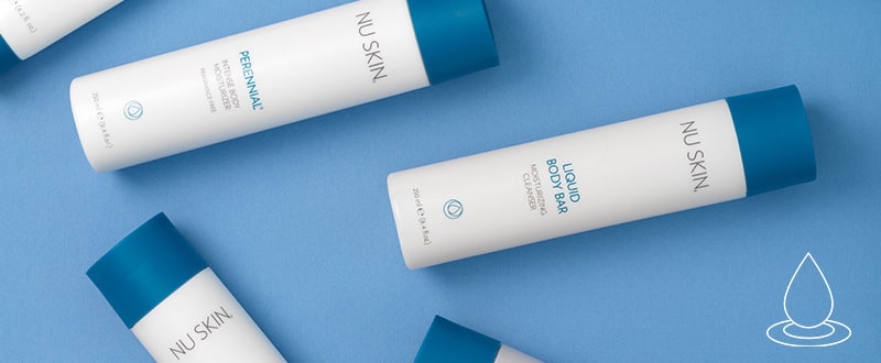 Verschiedene NuSkin Pflegeprodukte (unter anderem der Perennial Intense Body Moisturizer) liegen verstreut auf glatter blauer Oberfläche.