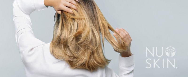 Frau mit weißer Bluse steht mit dem Rücken zur Kamera, sie kämmt sich mit den Händen durch ihr weiches dunkelblondes Haar. Bild wurde mit dem NuSkin Logo in der rechtne unteren Ecke gebrandet.