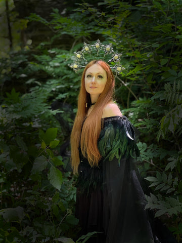 A redheaded woman in black stands among foliage and looks up at the sun.