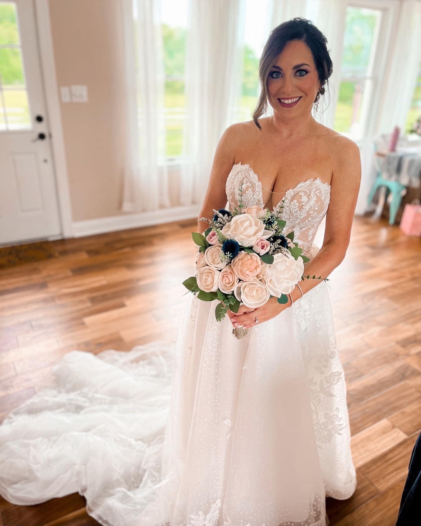 a bride standing in a room wearing a strapless wedding gown holding a bouquet of pink and white wood flowers