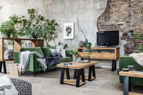 A living room with an industrial design. The living room has a rustic feel with a large exposed brick wall on the right side and a concrete wall on the left. There is a green sofa with various cushions and a throw blanket. In front of the sofa, there is a wooden coffee table with a plant, books, and a cup on it. There is also a small wooden side table with a plant on it. Above the coffee table, there are two framed pictures on the wall. The room has a cozy and rustic atmosphere.