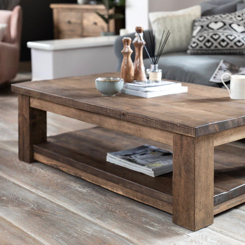 The main focus of the image is a wooden coffee table with a rustic finish. The coffee table is rectangular and has a lower shelf. On top of the coffee table, there are a few decorative items including a small stack of books, a bowl, two tall wooden figurines, and a glass container with reed diffusers. In the background, there is a glimpse of a beige armchair and a wooden sideboard or dresser. There is also a cushion with a geometric pattern on a piece of furniture next to the coffee table.