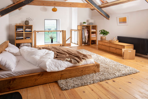 The bedroom has a rustic and cozy feel with wooden walls and ceiling. There is a bed with white bedding and a brown blanket. Next to the bed, there is a small wooden nightstand with a plant on it. The room has a sloped ceiling with a skylight window. There is also a rug on the wooden floor