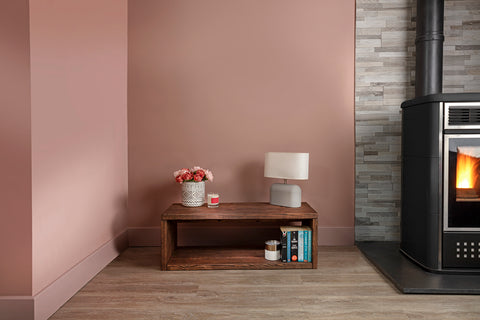 The image on the screen is of a cozy room setting. The room has a peach-colored wall on the left and a gray brick wall on the right. There is a wooden coffee table in the center of the room with a small vase of flowers, a lamp, and a few books on it. The floor has a checkered patterned rug. On the right side of the image, there is a modern wood-burning stove with a visible flame through its glass door