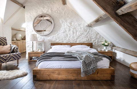 An image of a cosy bedroom. The bedroom has a rustic and warm feel with wooden elements. The bed is the focal point, with a wooden bed frame and white bedding. There's a grey patterned throw blanket on the bed. Above the bed, there is a round mirror with a wooden frame. The room has a slanted ceiling with wooden beams.