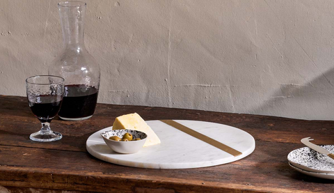 A white marble cheese board on a dark wooden table. There is a block of cheese on the board, and a glass of wine behind it.