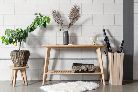 a wooden table against a white brick wall. On the table, there is a small potted plant with green leaves, a vase with two decorative palm leaves, and a wooden holder with pens. Behind the table, there is a small wooden stool with another potted plant.