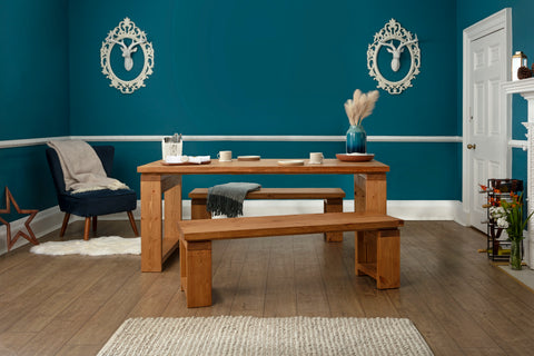A dining room with a teal-coloured wall. On the wall, there are two decorative deer heads that are painted white with an ornate frame. In front of the wall, there is a wooden dining table with a vase containing dried plants. To the left of the table. there is a chair with a grey cushion. The floor has a bobbled rug. There are matching wooden benches on each side of the dining table.