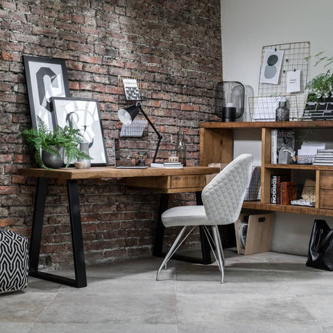 The picture shows an image of a cozy workspace. The workspace has a rustic feel with a brick wall in the background. There is a wooden desk against the brick wall with black metal legs. On the desk, there is a small black lamp and some decorative items. Leaning against the brick wall, there are a few picture frames. There is also a modern chair with a white and grey patterned upholstery in front of the desk. The floor has a checkered pattern.