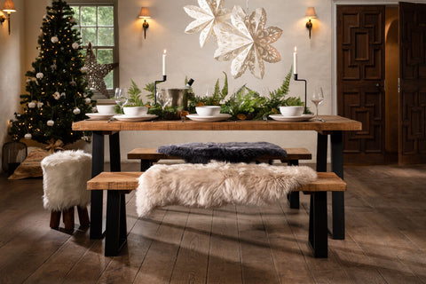 A wooden dining table that is set for Christmas dinner. The dining table has metal legs and two fur blankets on each bench on either side. To the left in the background is a lit up Christmas tree. The dining room is decorated for Christmas and giving a cosy atmosphere.