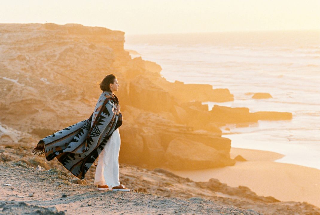 Person wrapped in a warm blanket, standing contemplatively on a coastal cliff at sunrise, symbolizing nurturing self-healing and the integration of profound shifts.
