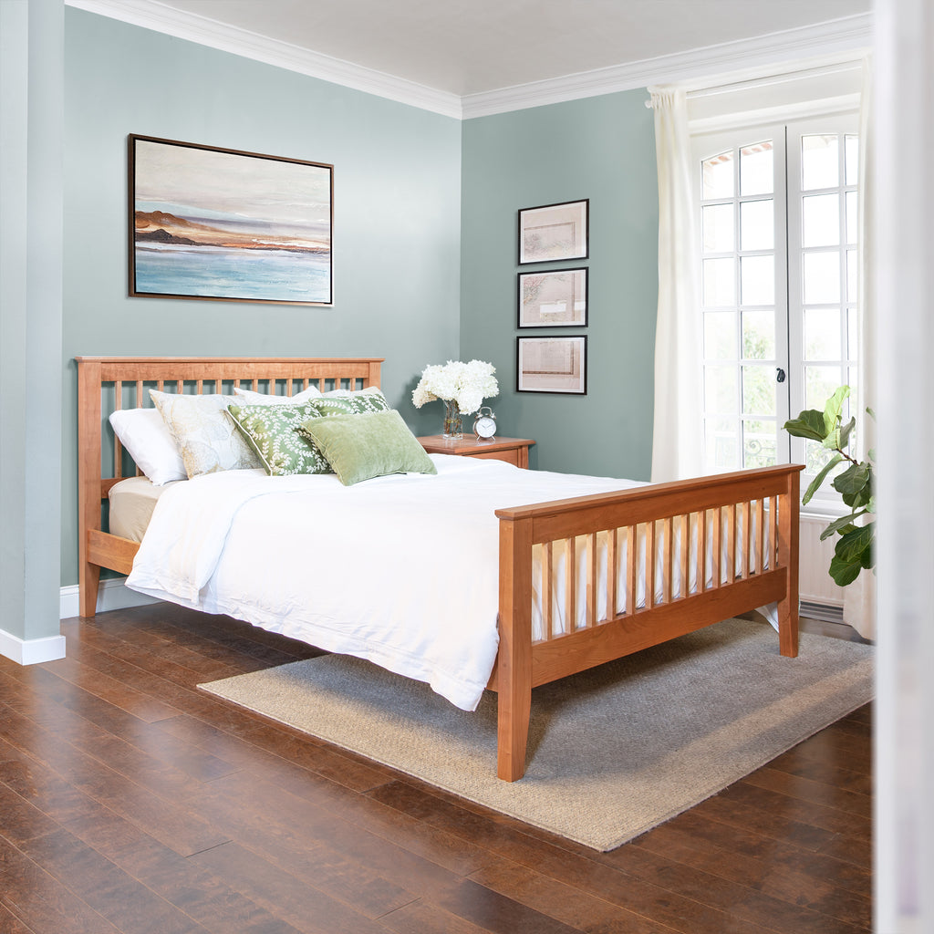 A cozy bedroom with light blue walls showcasing a Lyndon Furniture American Mission Bed frame, adorned with white bedding and green throw pillows. The room features a beige rug on wooden flooring, two framed pictures above the bed, another larger framed picture to the left, and a potted plant by the French doors. The stylish décor highlights high-quality American-made furniture from our collection.