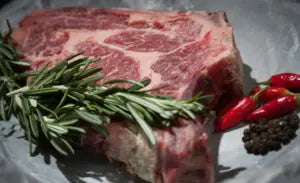 Raw steak with rosemary, red chili peppers, and black peppercorns on a plate.
