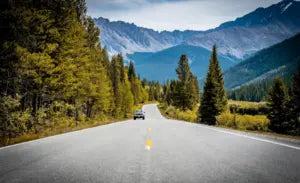 A car on a scenic mountain road surrounded by trees and distant peaks.
