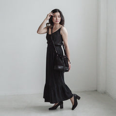 A woman wears a black drawstring bucket bag made from faux suede and cactus leather, a long black viscose dress and black heeled shoes