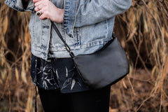 A model wears a small curvy black crossbody handbag made from cactus leather, with a blue denim jacket and black trousers