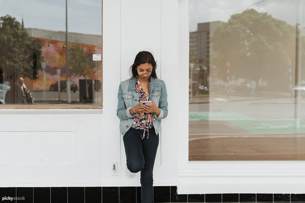 Young Indian female stands confidently leaning against a white shopfront window. She's on her phone. 