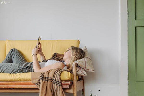 Lady lies reading kindle on a yellow retro sofa. There is a lime green door to the room closed, to the right of frame. 