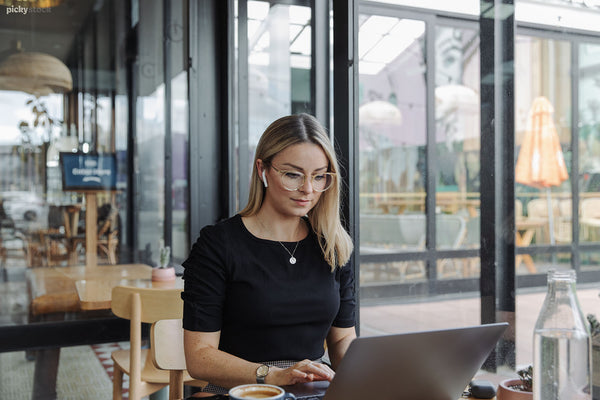 Blondie lady in black top with earpods in sits in a cafe, working on laptop. 