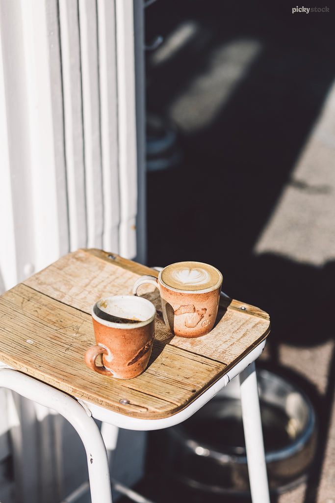 Two ceramic coffee mugs sitting on a stool outside a cafe. 