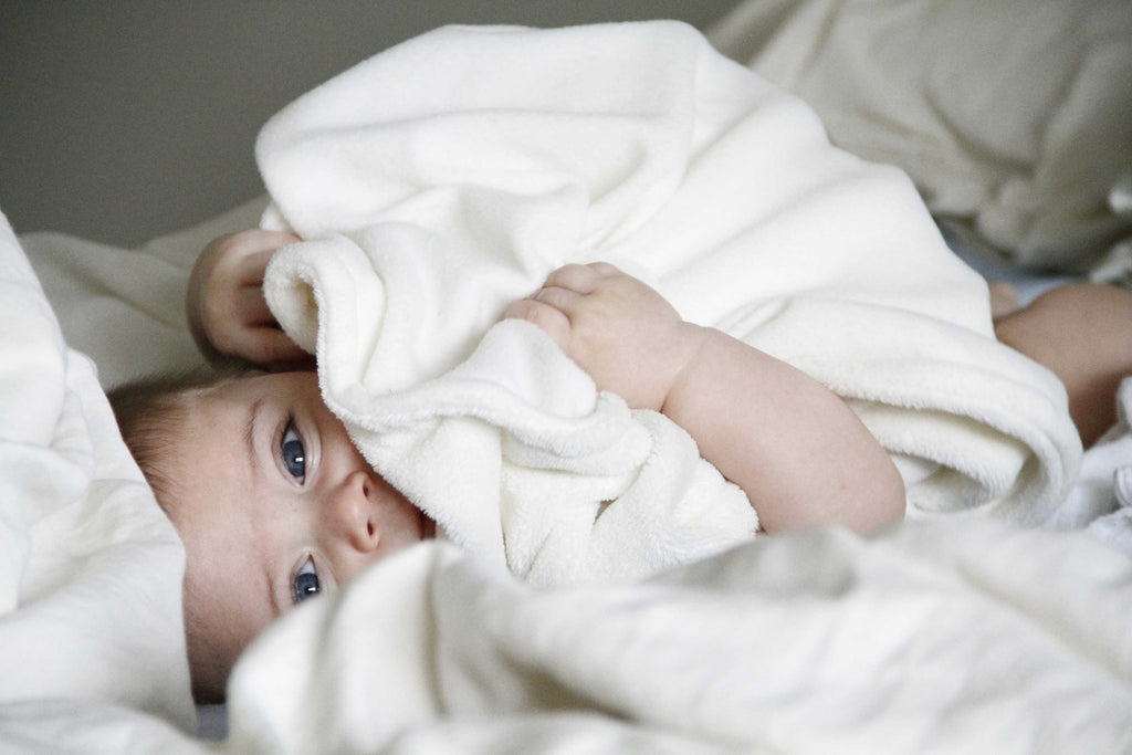 A serene baby peeking out from under a white blanket, with captivating blue eyes that suggest curiosity and wonder, nestled in a soft bed for a peaceful rest