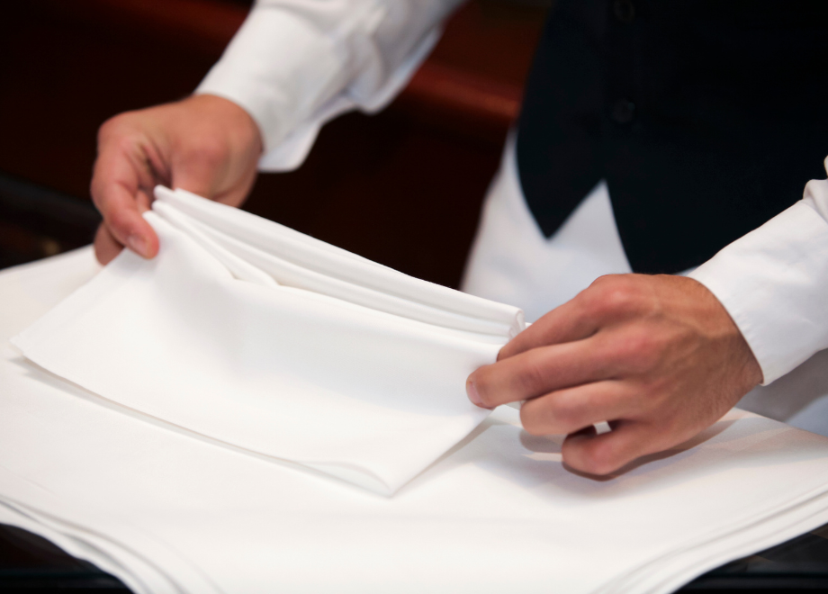 A person in a white dress shirt and black vest is folding white napkins