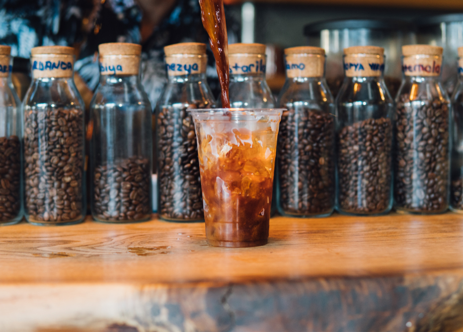 A clear plastic cup of cold brew is being filled at a bar. There are jars of coffee beans in the background