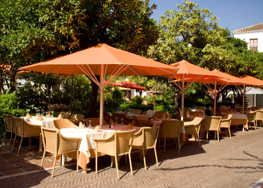 an outdoor dining set up with multiple tables, and wooden/wicker chairs. there are orange and white tablecloths and orange umbrellas. for each table. it is a sunny day out, and there is a row of trees running parallel to the dining area