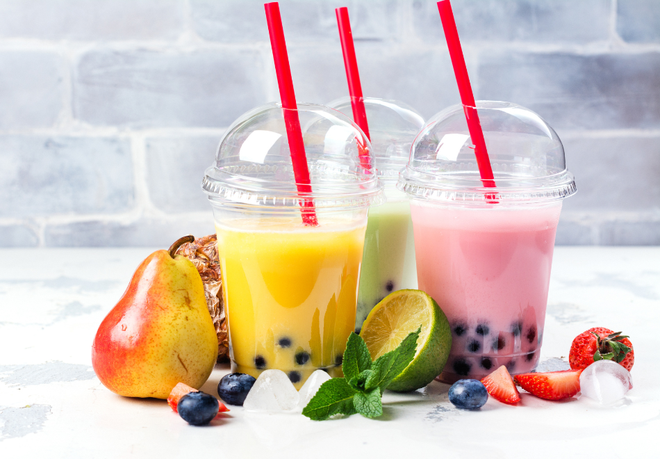 three colorful smoothies with clear plastic dome lids on a table surrounded by fresh fruit