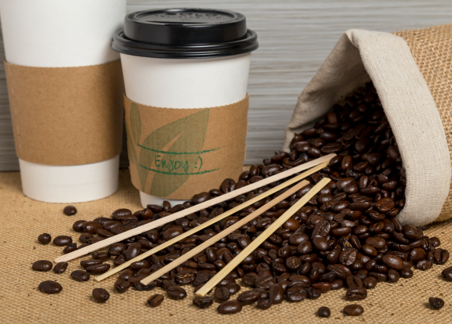 spilled coffee beans with a variety of stirrers on top and paper cups behind