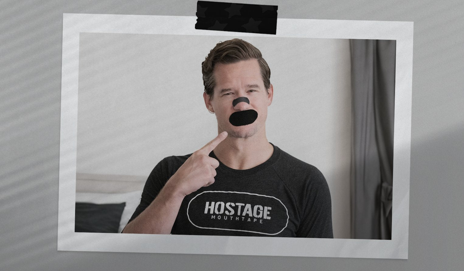 Man with tape over mouth pointing at it, wearing a shirt labeled 'HOSTAGE MOUTHTAPE'.