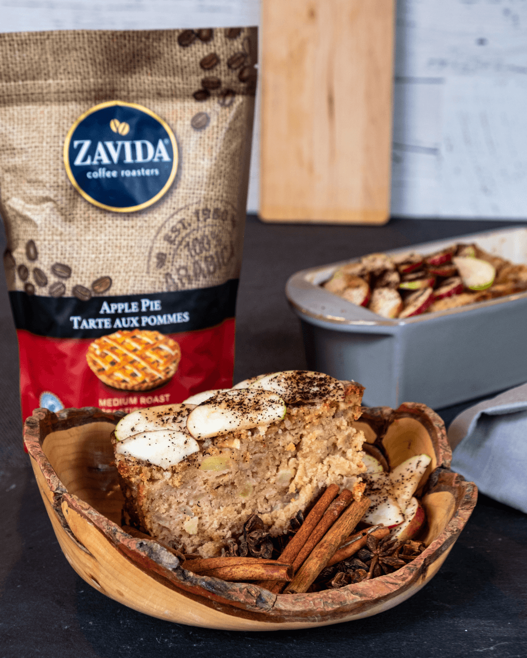 Easy apple coffee cake is cut and placed in a wooden bowl next to the pan and a bag of Zavida's Apple Pie Coffee
