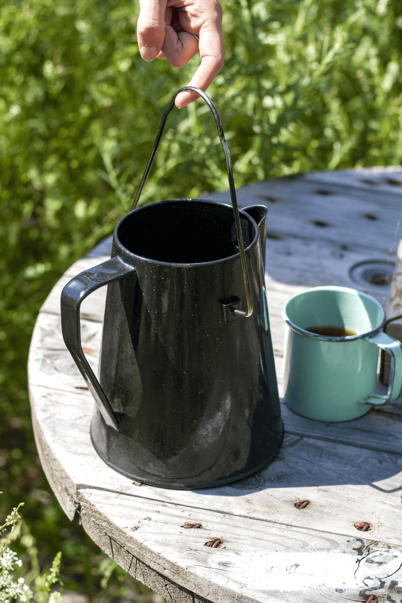 Cowboy coffee pot sits on a table outdoors beside a half full coffee mug.