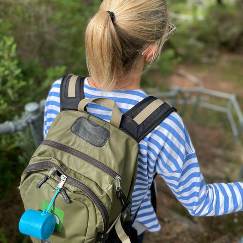 Woman with a backpack on her back leaning on a rail outdoors in the Australian bush. There is a blue Solmate refillable sunscreen applicator attached to the backpack.