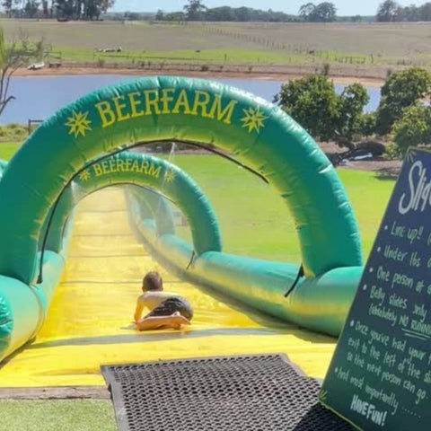 boy sliding down a large water slide outdoors with a dam and bush in the background and a pub menu in the left hand corner