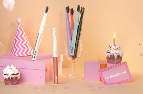 All variants of Perfora's Electric Toothbrushes in a cup and two metallic brushes outside the glass with a cupcake on the side