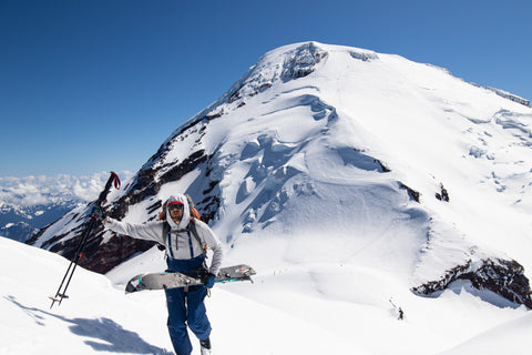 a snowboarder hikes up Mount Sherman with Mount Baker in the background