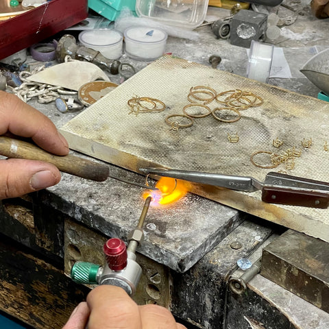 Two hands at a jeweler's workbench: one is holding a lit torch, the other a tool.  The jeweler is soldering.