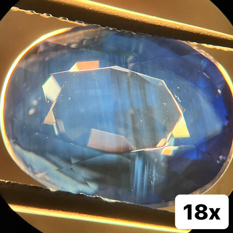 A natural blue sapphire under 18x magnification, displaying straight line color zoning and cloudy rutile inclusions.